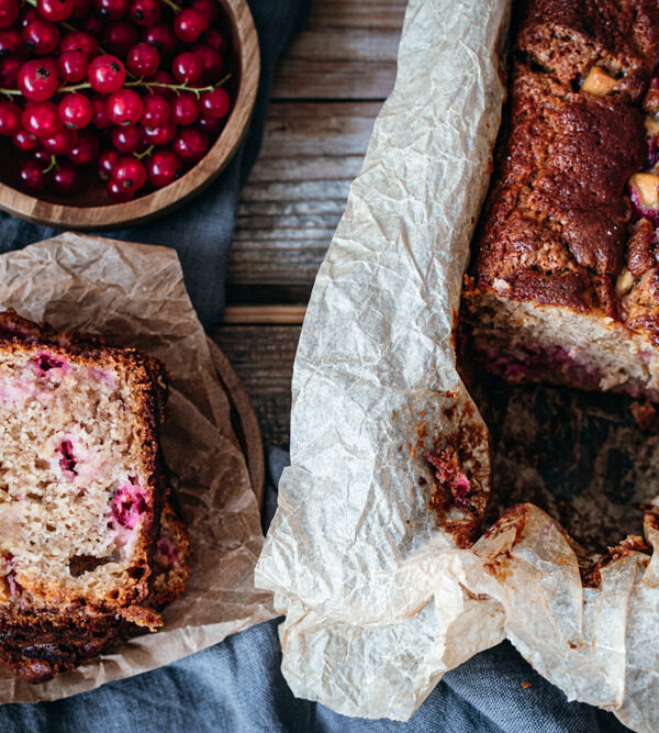 Flatlay eines Bananenbrotes mit Roten Johannisbeeren, links oben einenyholzschale mit den roten, kleinen Beeren, links unten ein Stapel aufgeschnittener Stücke und rechts das restliche Brot in einer Kastenform mit Backpapier