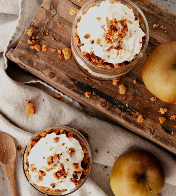 zwei Gläser mit Schichtdessert mit Schlagsahne, zwei Äpfeln, einer Holzkiste und einem beigen, groben Tuch