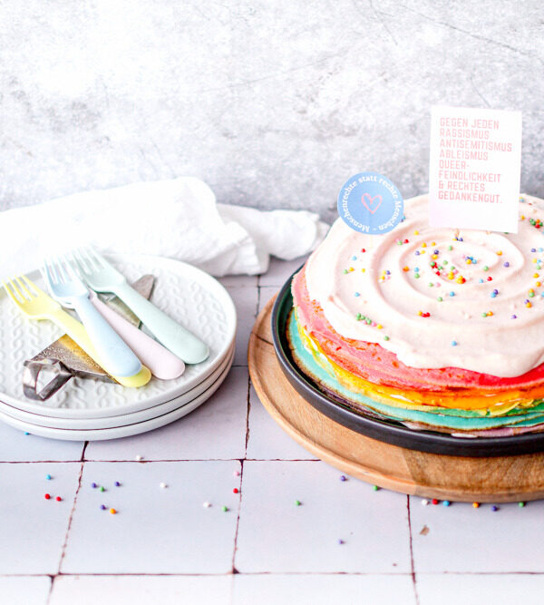 Links ein Kuchen aus gestapelten Regenbogen-farbigen Pfannkuchen mit rosé Creme on top und Demo-Schildern gegen rechts geschmückt. Links ein Stapel aus drei grauen Tellernden mit farbigen Kuchengabeln darauf