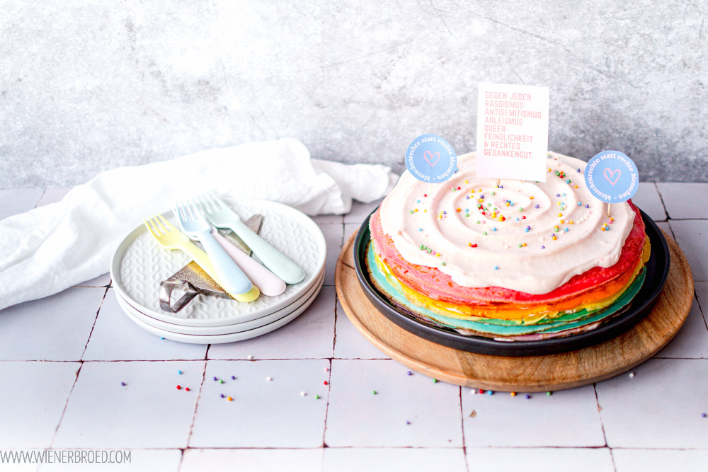 Links ein Kuchen aus gestapelten Regenbogen-farbigen Pfannkuchen mit rosé Creme on top und Demo-Schildern gegen rechts geschmückt. Links ein Stapel aus drei grauen Tellernden mit farbigen Kuchengabeln darauf