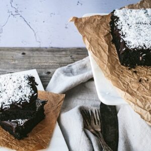 Schwedischer Schokokuchen mit Kokoraspeln, links zwei Stücke übereinander, rechts geschnittene Stücke auf einer weißen Platte auf Backpapier