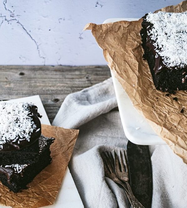 Schwedischer Schokokuchen mit Kokoraspeln, links zwei Stücke übereinander, rechts geschnittene Stücke auf einer weißen Platte auf Backpapier