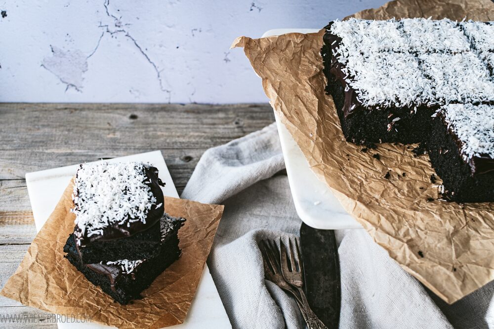 Schwedischer Schokokuchen mit Kokoraspeln, links zwei Stücke übereinander, rechts geschnittene Stücke auf einer weißen Platte auf Backpapier