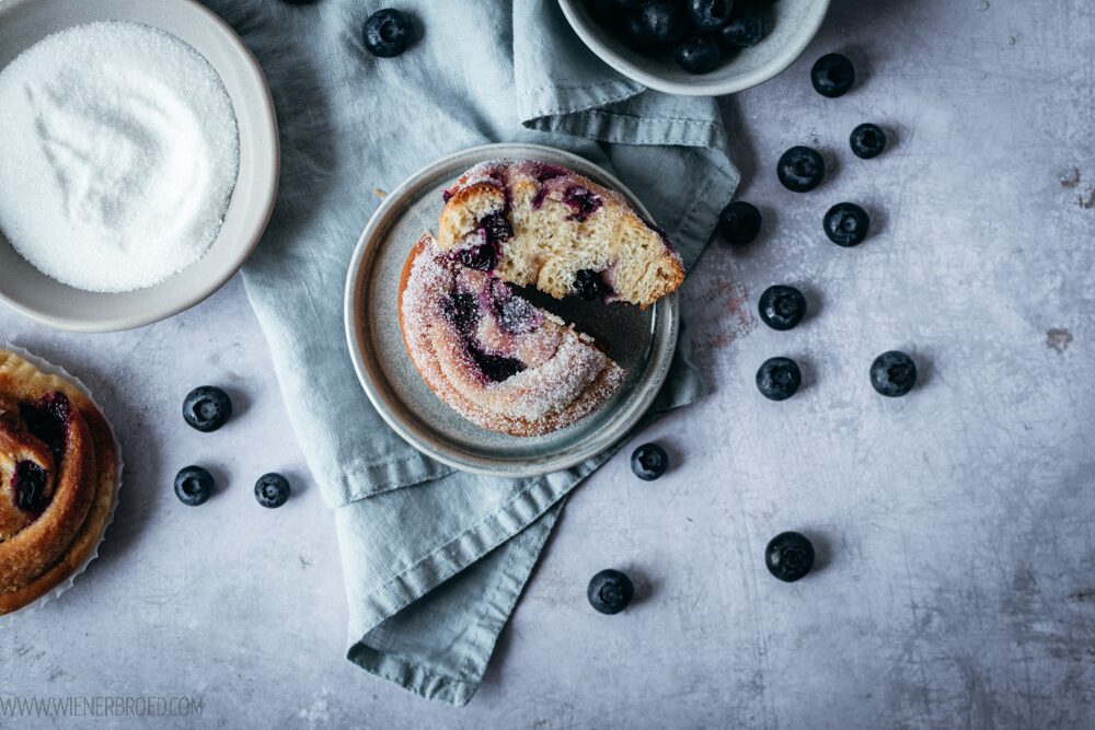 Blick auf eine aufgeschnittene Hefeschnecke mit Blaubeeren und Zitronenbutter, links eine Schale mit Zucker, rechts frische Blaubeeren