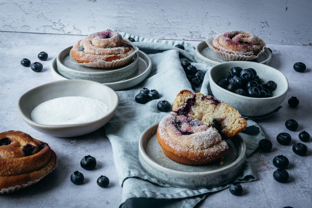 Blick auf einen Tisch mit 4 Blaubeerschnecken aus  Hefeteig, einer Schale mit weißem Zucker und rechts ein Schälchen mit Blaubeeren